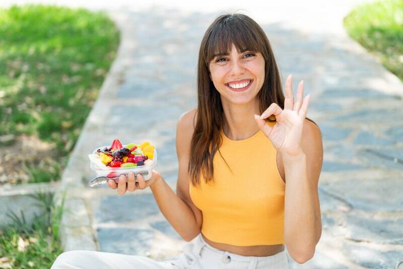 Young woman holding a bowl of fruit at outdoors showing ok sign with fingers Young woman holding a bowl of fruit at outdoors showing ok sign with fingers